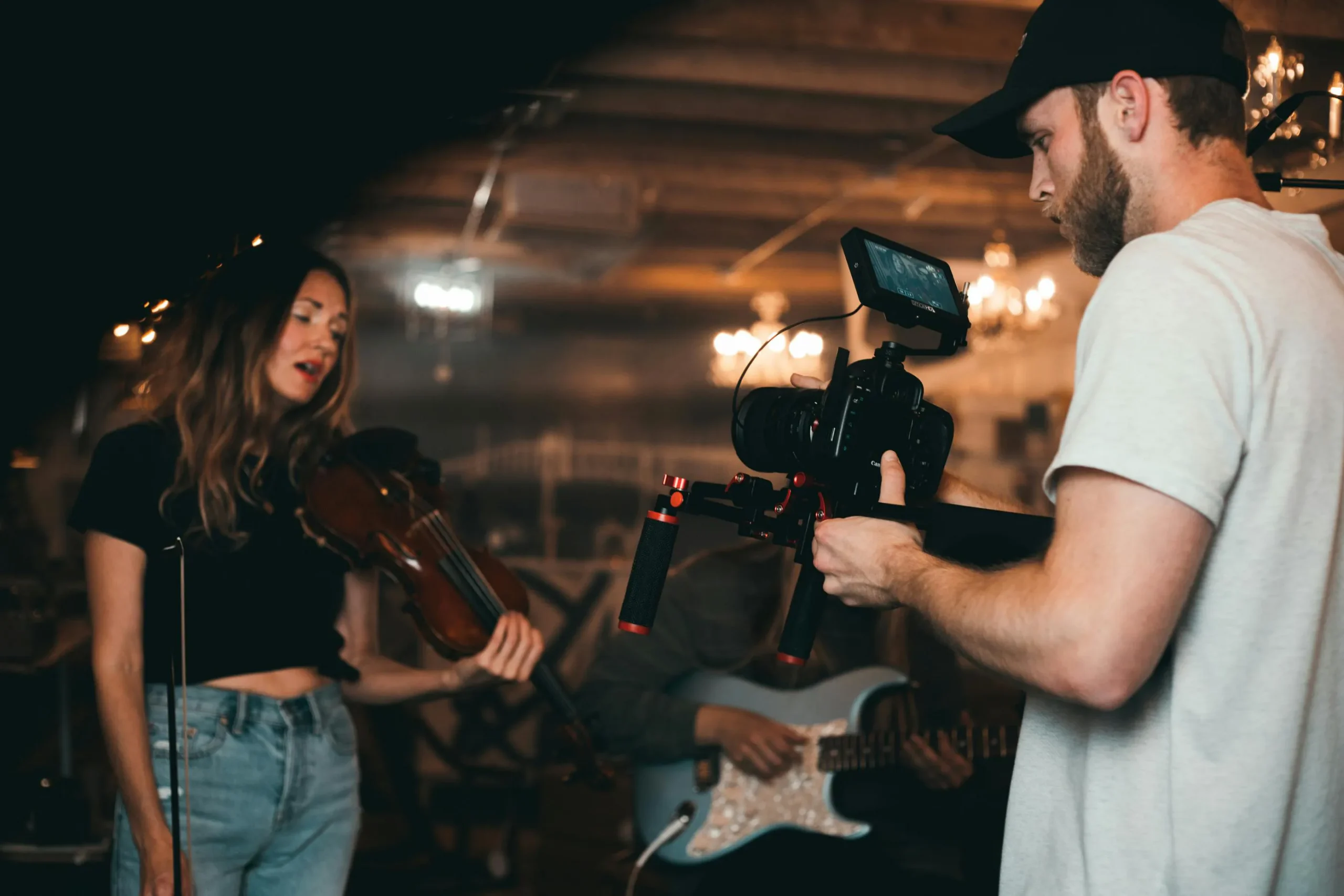 Cinematographer filming a musician playing violin and another playing guitar in a warm, ambient studio environment