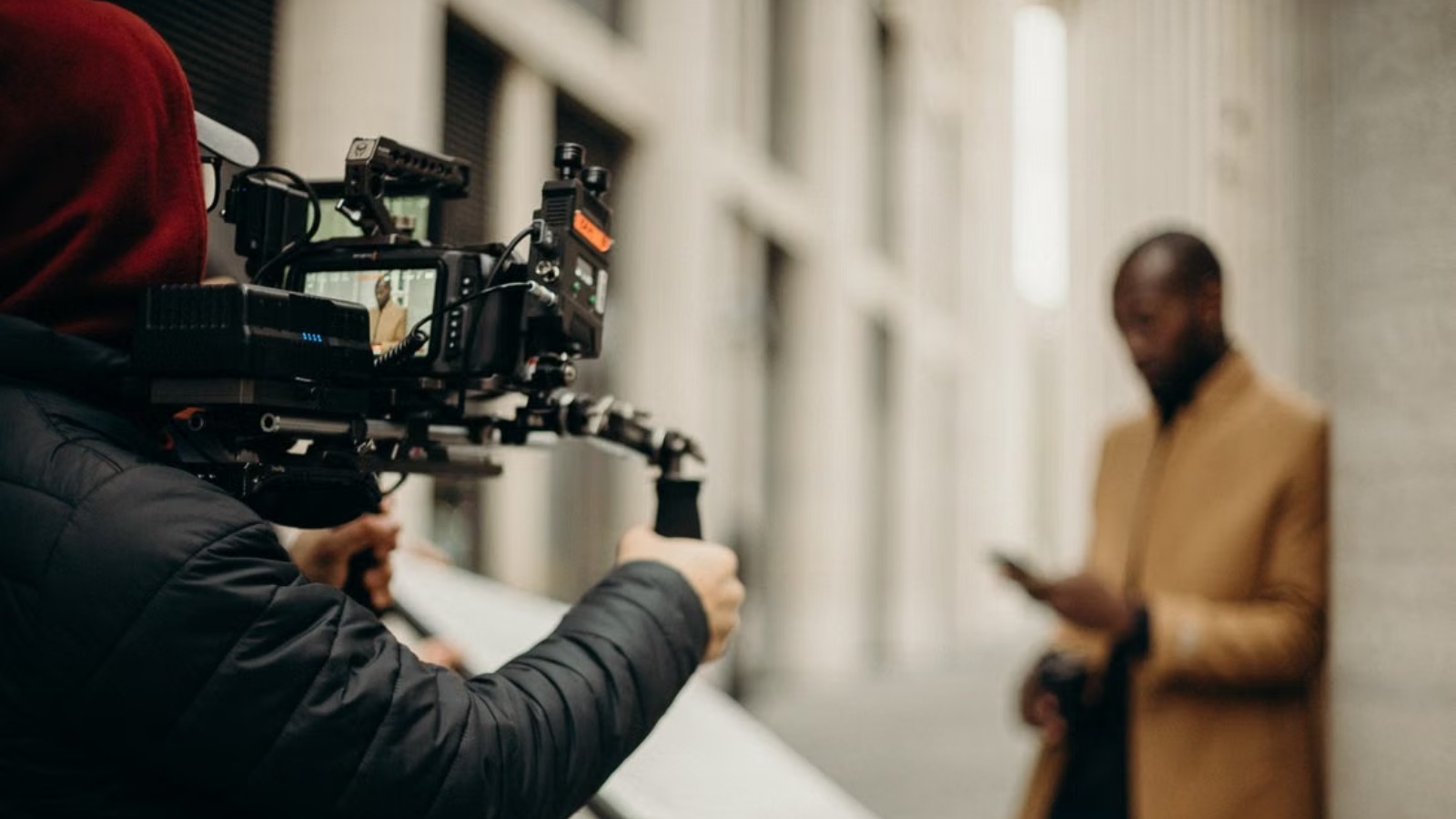A camera operator holds a professional rig, filming a man in a tan coat